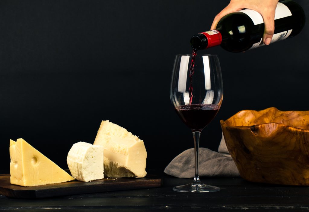 Large chunks of cheese sit on a wooden board, while a hand out of the shot pours a large glass of red wine which students can experience on a food and wine tour of Switzerland. This image is used in the Students Fare student travel blog, "Spring Break in Switzerland," which is a travel guide to celebrating spring vacation in the Swiss Alps.