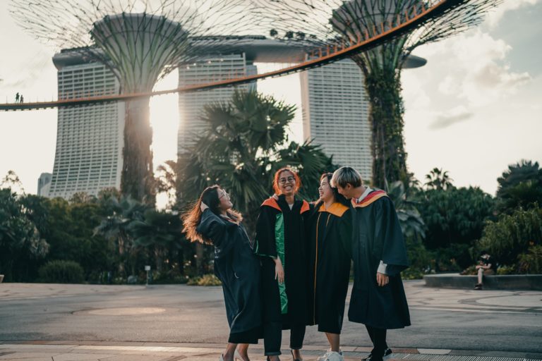 A group of four university students in their graduation gowns celebrating under beautiful architecture. The featured image in the Students Fare student travel blog, "Best College Towns in America," which describes the top university cities in the USA.