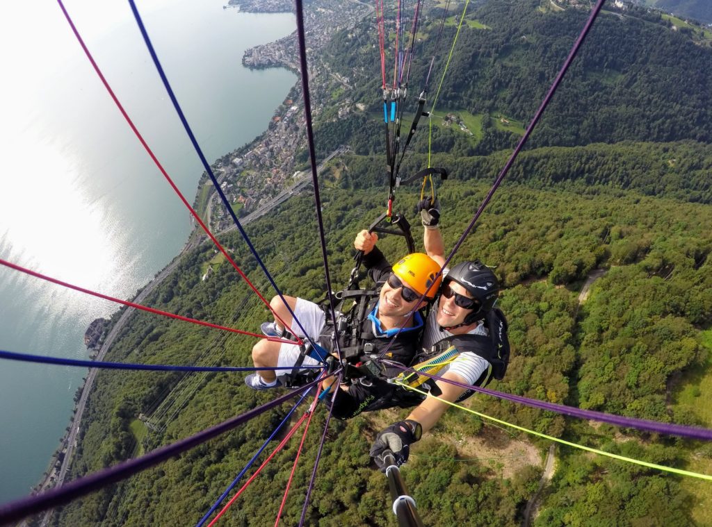 A birds-eye image of a paraglider and instructor flying high above green hills and enjoying a paragliding tour! This image is used in the Students Fare student travel blog, "Best Paragliding Destinations," which is a travel guide for paragliding all around the world.