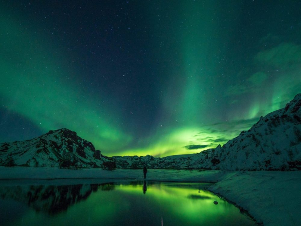 A lone student traveler standing beside the lake admiring the Northern Lights during a tour in Thórsmörk, Iceland.