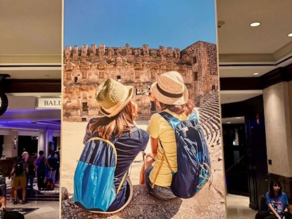 Advertisement banner of two people sitting at the 2024 SYTA event for Students Fare to discuss flights, tours, hotels, and academic pursuits.